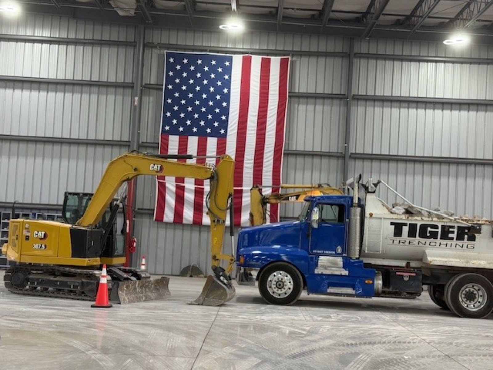 Blue Tiger Trenching dump truck and CAT excavator parked in the shop beneath an American flag